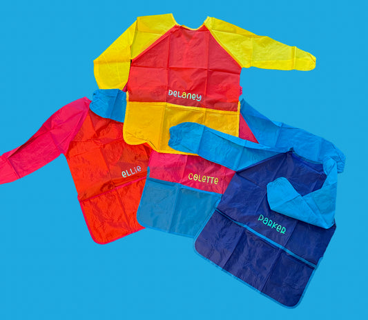 Custom Artist Smock for kids featuring a protective waterproof design, personalized with the child’s name. Ideal for pint-sized Picassos and little artists, this lightweight and durable plastic apron protects clothing from paint and glue. It includes a big front pocket for paint supplies and comes in various colors and font styles.