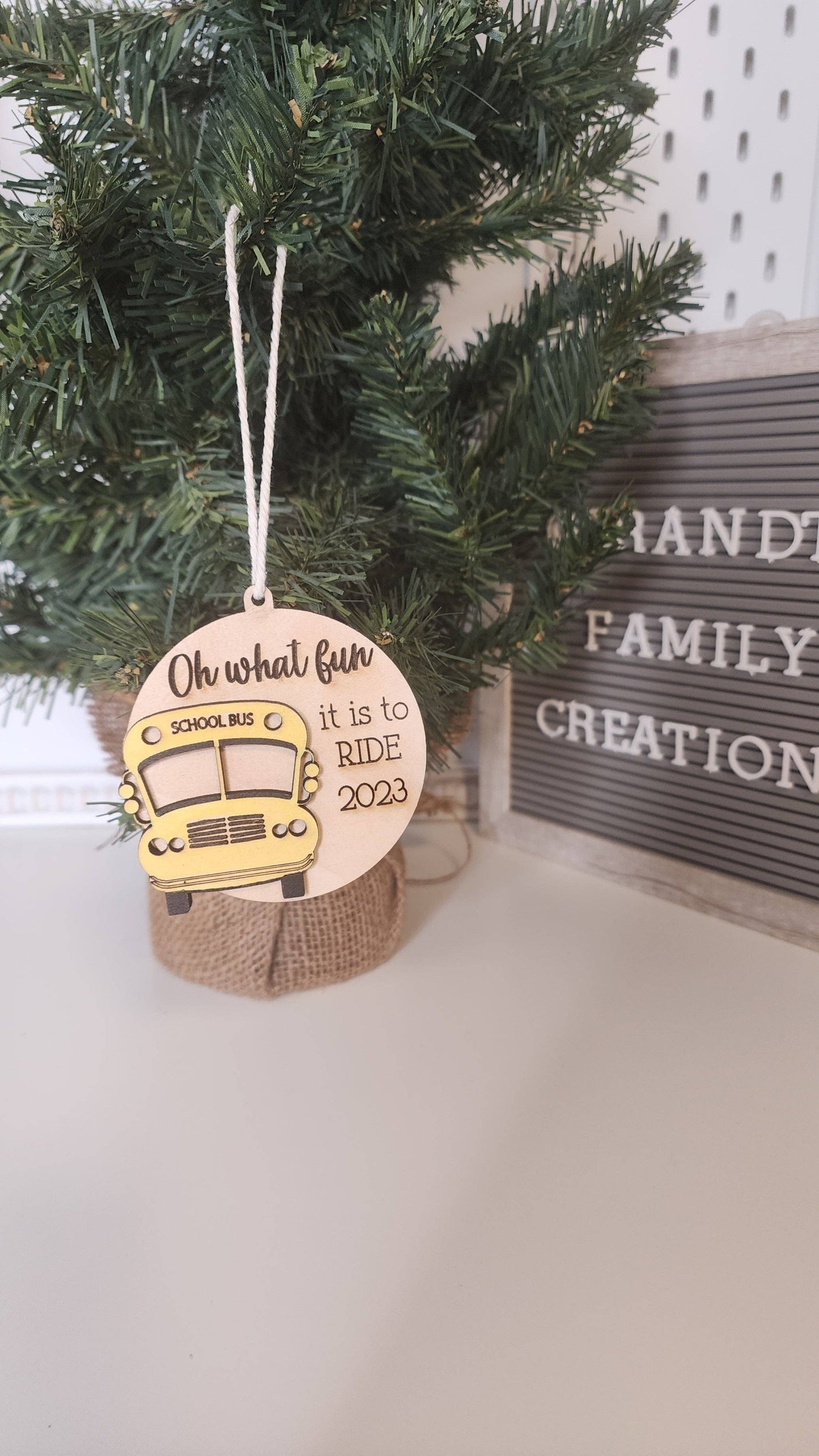 A festive Christmas tree ornament featuring a wooden design with a yellow school bus and the text 'Oh what fun it is to ride 2023.' The ornament hangs on a small Christmas tree with a burlap base, adding a personalized touch to holiday decor.