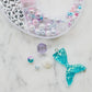 Aquamarine mermaid jewelry kit featuring alphabet beads, shell beads, iridescent star and heart beads, a mermaid tail charm, and pink and clear string. Perfect for creating your own bracelets or necklaces. Made by women-owned small STACKS. Warning: small parts.