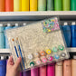 Coloring book with markers held in front of a shelf of colorful paint bottles.