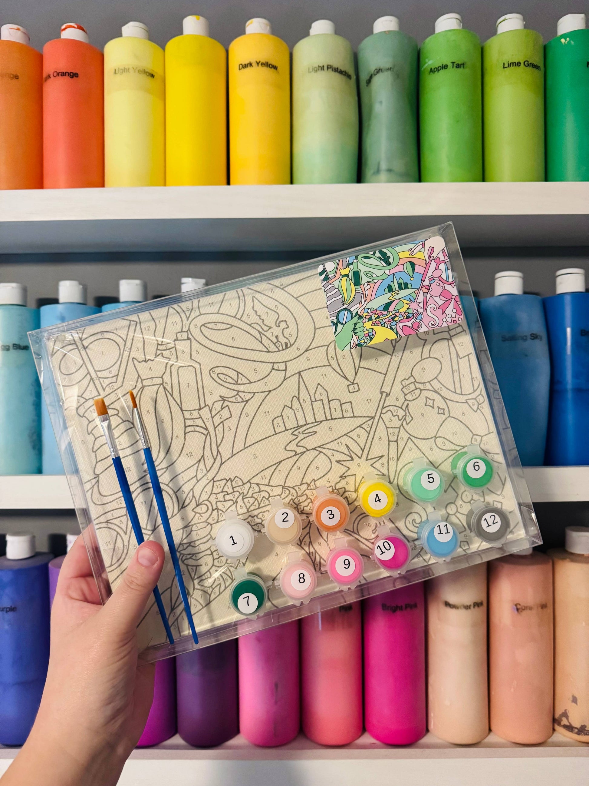 Coloring book with markers held in front of a shelf of colorful paint bottles.