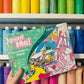 Colorful 'Paint Me!' paint by number kit held in front of a shelf with paint bottles.
