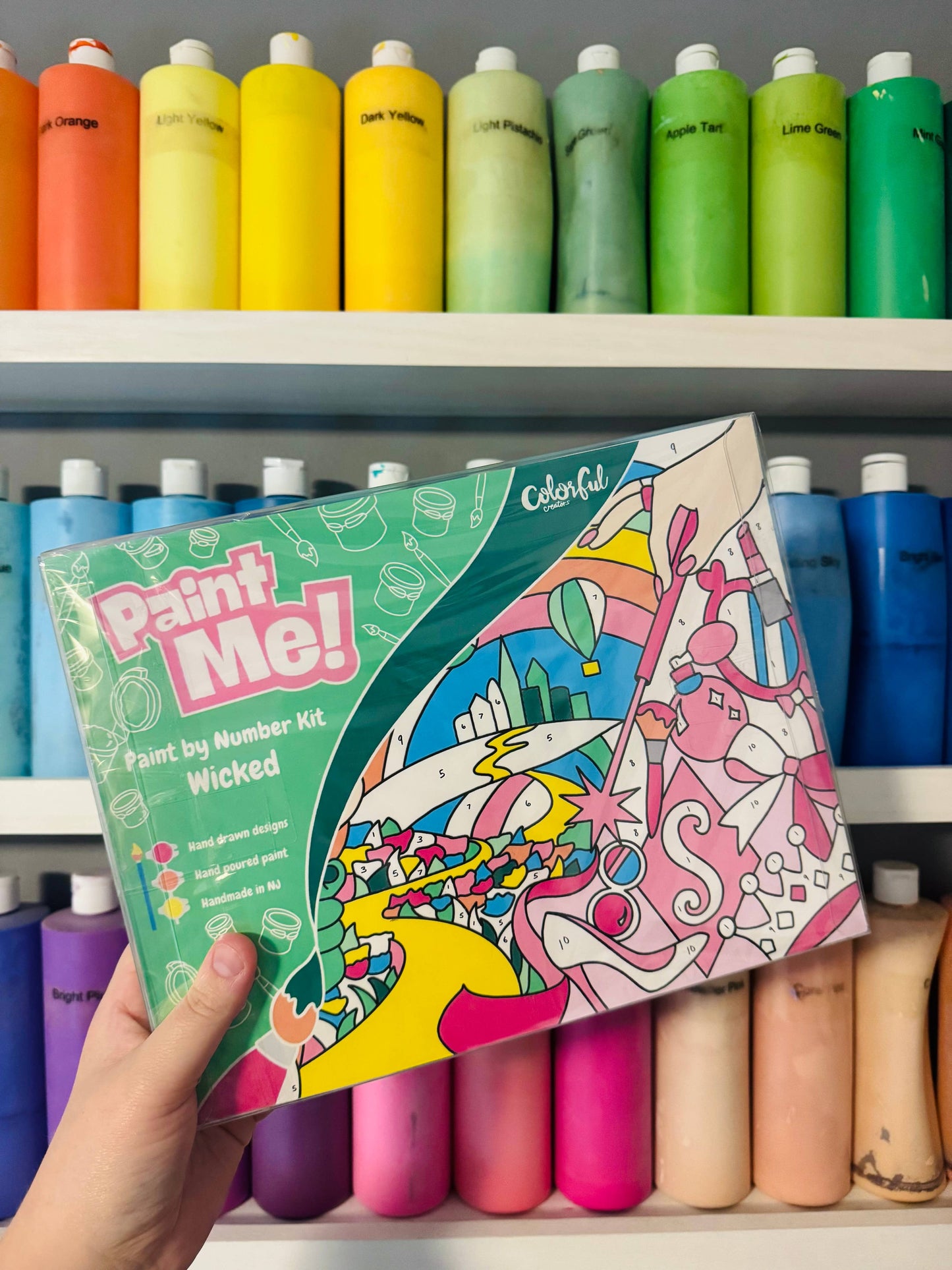 Colorful 'Paint Me!' paint by number kit held in front of a shelf with paint bottles.