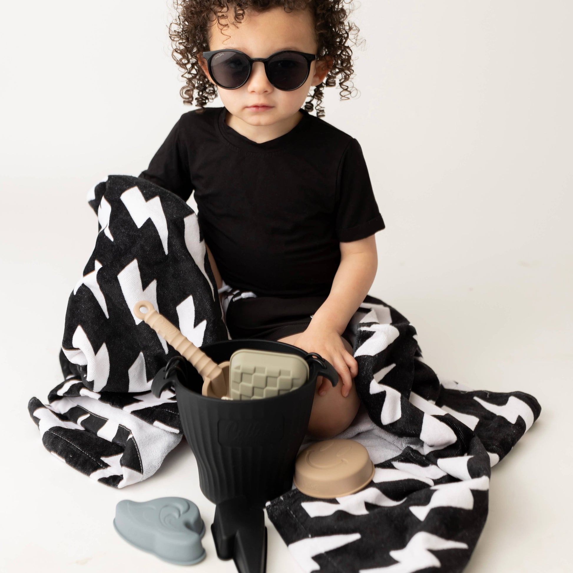 Child wearing sunglasses sitting on a black and white patterned blanket with toys around