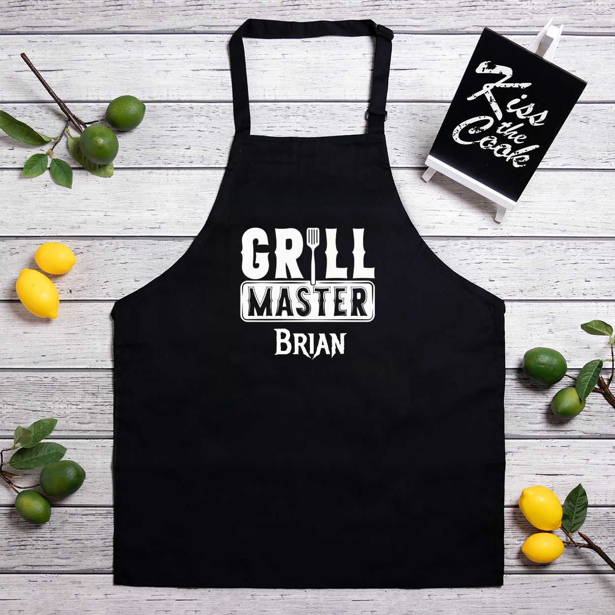 Personalized black kitchen apron with slider neck adjustment for easy sizing. Made from a 7.5 oz poly-cotton blend, featuring a one-sided 10.25"x10.25" design. Machine-washable for convenience, perfect for chefs and home cooks. Add your name for customization.
